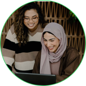 Two women, one wearing a headscarf, looking at a laptop, smiling.